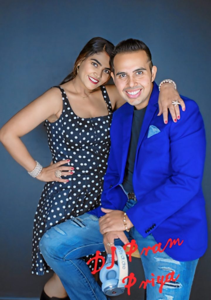 The image features an older picture of DJ Pram and MC Priya, showcasing their friendly and vibrant connection. DJ Pram is dressed in a stylish blue blazer, exuding confidence, while MC Priya stands beside him in a chic polka dot dress, radiating warmth and elegance. Both are smiling brightly, embodying a lively and approachable aura. The simple background ensures the focus remains on their engaging personalities and the camaraderie they share.