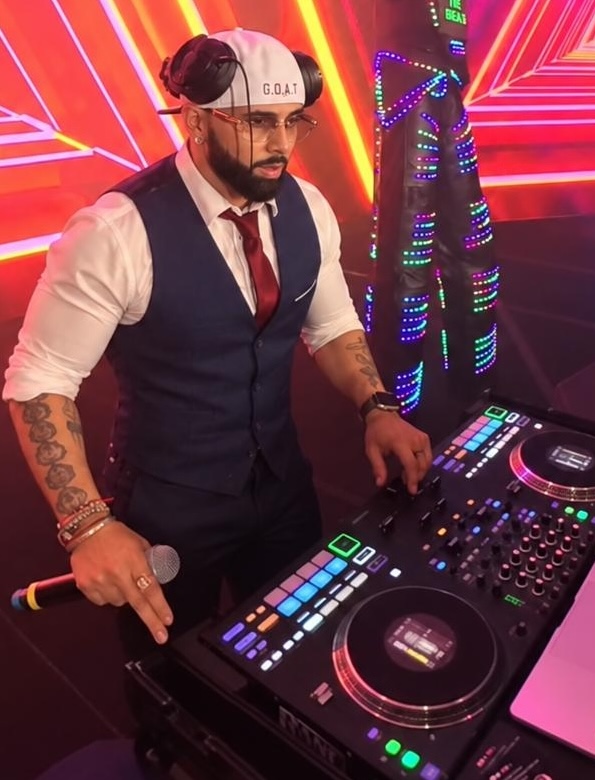 The image showcases DJ Pram, an Indian DJ renowned for his work across Orlando, Tampa, Miami, Florida, and Georgia, standing above a set of turntables. He is captured in a dynamic pose, pointing towards the camera with an engaging expression. The lighting creates a striking contrast with warm and cool tones, highlighting his focused demeanor. His cap and casual attire add a stylish touch, while the background subtly fades, keeping the focus on his commanding presence at the DJ booth.